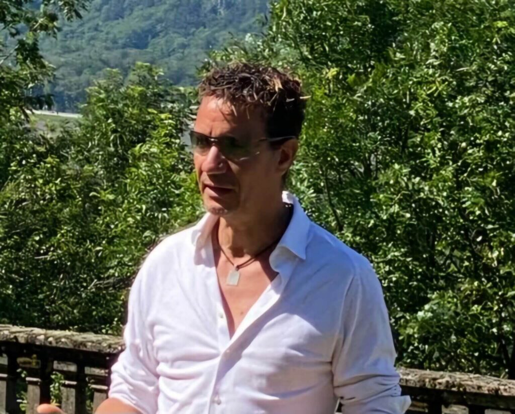 Abdel Anik en buste avec chemise blanche entre ouverte sur un fond de verdure ensolleillé